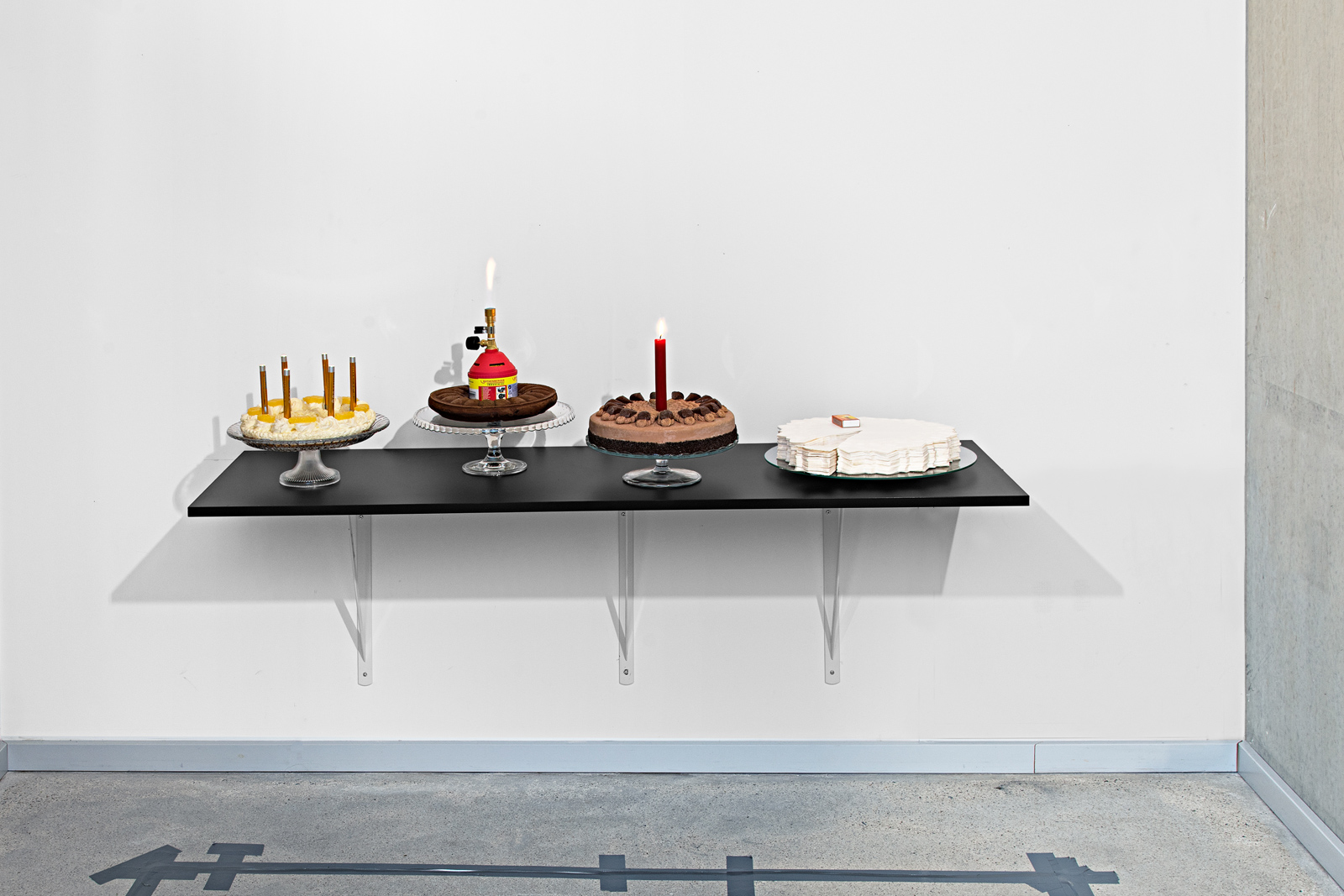 Kunst-Objekt von Josephine Riemann: Echte oder künstliche Kuchen sind zusammen mit Diagnostiktaschenlampen, einer Kerze, einem Bunsenbrenner oder Streichhölzern ausgestellt.  